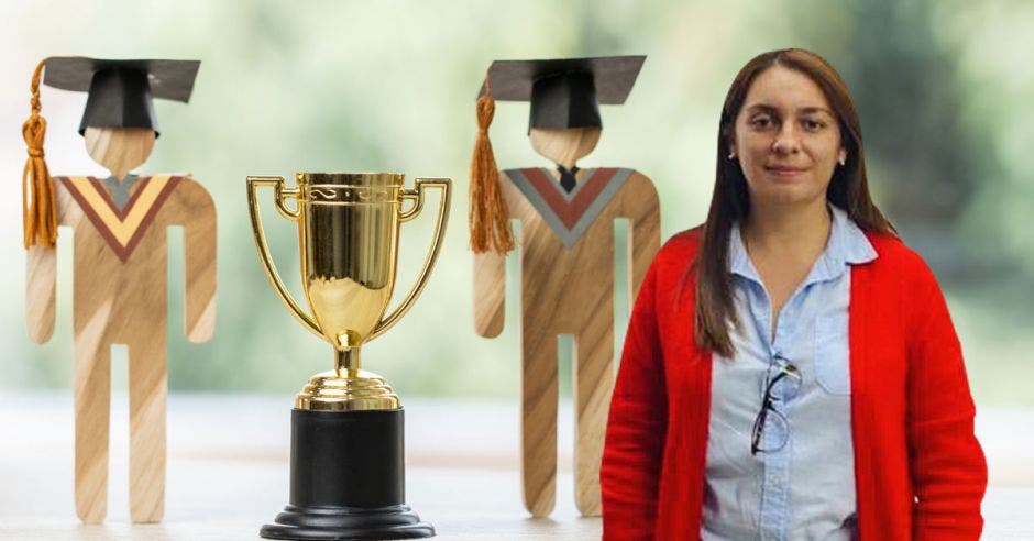 mujer con sueter rojo y camisa azul de fondo muñecos de madera con birretes y en medio un trofeo dorado